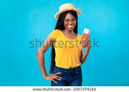 Cheap Flight Tickets. Cheerful Black Lady Tourist Holding Boarding Pass And Passport Posing Over Blue Studio Background, Wearing Hat And Sunglasses. Summer Vacation, Tourism And Traveling