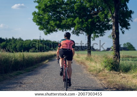 Photo from behind of a woman cyclist, riding a road bike on a road in the middle of nature. Wearing a pink and green t-shirt Royalty-Free Stock Photo #2007303455