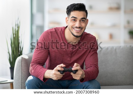 Emotional middle-eastern guy playing video game with joystick at home, copy space. Happy millennial arab man enjoying weekend, sitting on couch in living room with joystick, gaming Royalty-Free Stock Photo #2007308960
