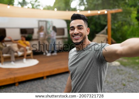 Young Arab guy taking selfie near motorhome, smiling at camera, resting on campsite with multiracial friends, creating happy memories, enjoying summer vacation outdoors, free space