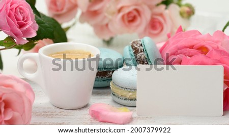 blank white business card and cup with espresso coffee and white ceramic cup with coffee and blue macaron on a white table, behind a bouquet of pink roses