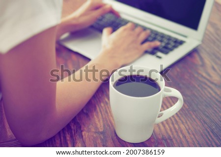 Work from home theme white cup with black coffee and blurred female hands using laptop on old dark wood texture with natural patterns table background. Technology's impact on people's live theme.