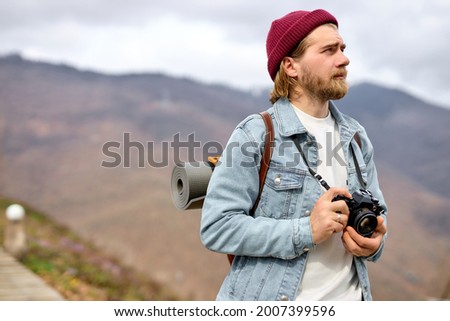 Handsome Caucasian man with backpack and camera is walking towards distant mountain. Bearded Man hiker hiking in mountains in spring. Traveler travels on country road, ecotourism, hiking.