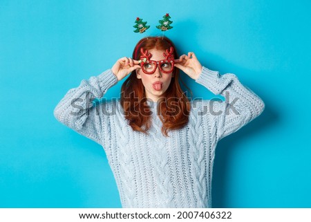Christmas party and celebration concept. Silly redhead girl enjoying New Year, wearing funny glasses and headband, showing tongue and staring left at logo, blue background
