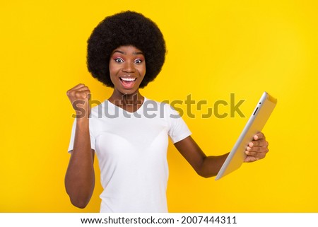 Photo of lucky funny dark skin woman dressed white t-shirt smiling rising fist holding modern device isolated yellow color background