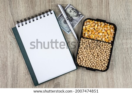 Top view mockup of open blank notebook on spiral, automatic pen. 100 dollar banknotes and samples of soybean and corn seeds, conceptualizing agricultural commodities. on a wooden table