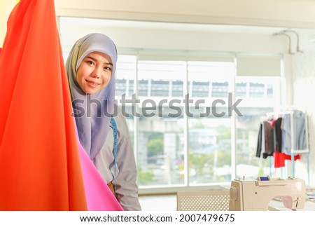Portrait of beautiful smiling asian muslim woman designer with hijab looking to camera in her home working studio room with orange and pink cloth, tape measurement and sewing machine.