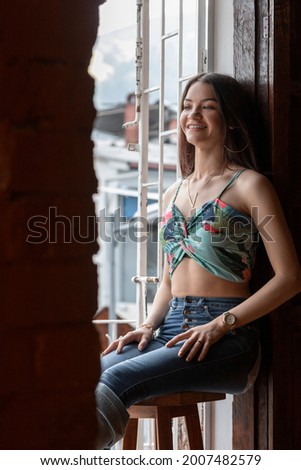 cheerful slim young woman in casual clothes sitting on a wooden bench next to a window in the daytime, wearing top and jeans, and looking outside, lifestyle