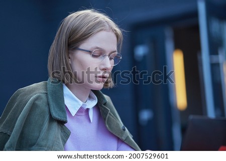 Young woman freelancer in glasses sits on cafe terrace at table with black laptop and types under bright sunlight. Female student distant learning foreign languages and working distantly. Royalty-Free Stock Photo #2007529601