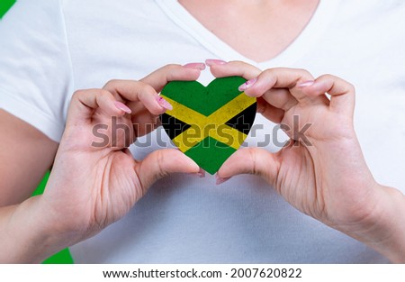 Woman in white t-shirt holds a heart in the form flag Jamaica on her chest. Photo for a postcard on Flag Day, Independence Day, travel, patriotism, other holiday