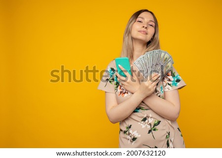 Photo of pleased happy emotional young woman posing isolated over yellow wall background using mobile phone holding money.