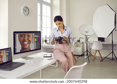 Photographer sitting on edge of table and looking at photos on her DSLR camera. Focused serious young woman working with post processing editing software on modern desktop, laptop and tablet computers