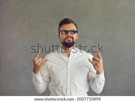 Self assured man wearing funny sunglasses. Studio shot of smirking smug looking student or businessman in white shirt and thug life glasses doing rock sign gesture standing isolated on grey background