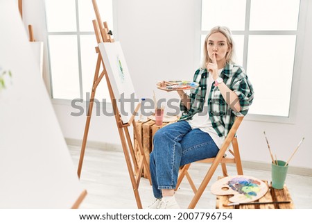 Young artist woman painting on canvas at art studio asking to be quiet with finger on lips. silence and secret concept. 