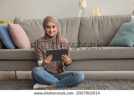 Funny video, online call, mobile app, meeting remotely, good offer, ad during covid-19 pandemic. Smiling arab female in hijab watching lesson on tablet in living room interior on floor, copy space