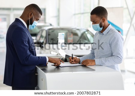 Black Man In Medical Mask Buying New Car In Dealership Office During Coronavirus Pandemic, African American Male Customer Signing Papers With Salesman In Modern Automobile Showroom, Free Space