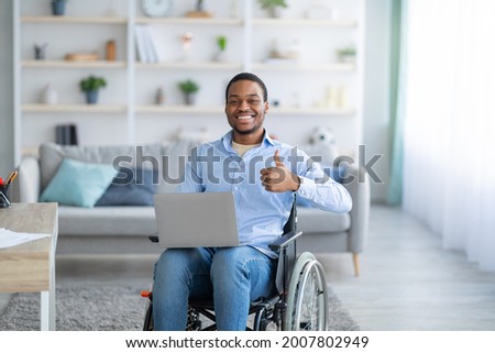Portrait of happy handicapped black guy with laptop showing thumb up gesture, smiling at camera indoors. Paraplegic young man in wheelchair recommending online job during covid pandemic Royalty-Free Stock Photo #2007802949