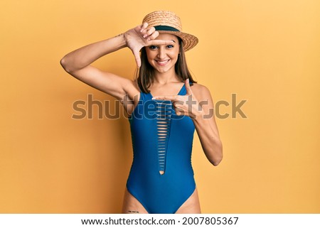 Young brunette girl wearing swimsuit and summer hat smiling making frame with hands and fingers with happy face. creativity and photography concept. 