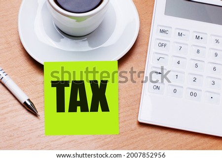 The wooden table has a white cup of coffee, a pen, a white calculator, and a green sticker with the text TAX. Business concept