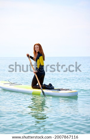 Caucasian red-haired woman with long hair in black wetsuit working out with stand up paddle board on water or in open sea, slim beautiful lady is engaged in water sport, extreme sport
