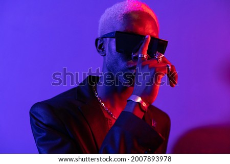 Portrait photo of a dark-skinned guy with a beard. Dressed in a black jacket and sunglasses. With metal ornaments on his hands Purple and pink light