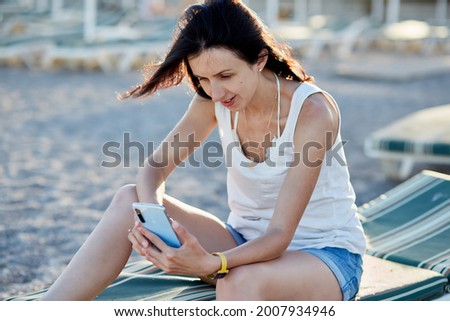 Young brunette with a phone on a sunbed on the beach near the sea. Vacation concept, taking a selfie, chatting with friends or freelancer, working remotely, webinar, communication, learning concept. 