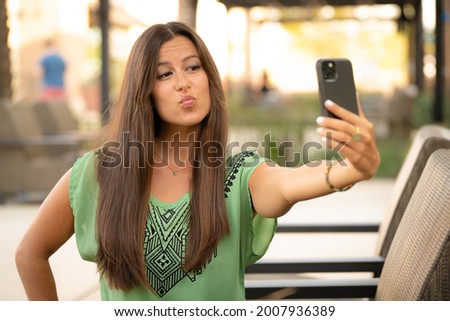 Beautiful Pretty Young Brunette Woman Taking Smiling Kissing Duck Face Funny Selfie Photo Picture with Phone in Natural Soft Daylight at Mall Café Outside