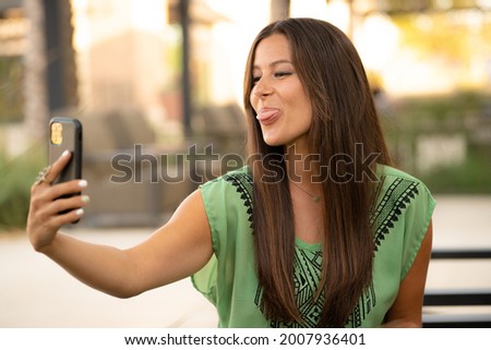 Beautiful Pretty Young Brunette Woman Taking Funny Selfie Photo Picture Sticking Tongue Out with Phone in Natural Soft Daylight at Mall Café Outside
