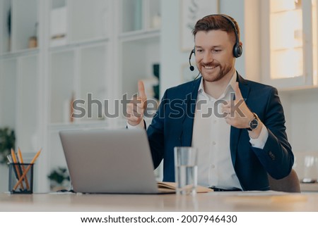 Happy male employee makes okay like gesture keeps thumbs up satisfied with colleague business presentation makes video call uses headphones with microphone wears formal clothing. Executive worker