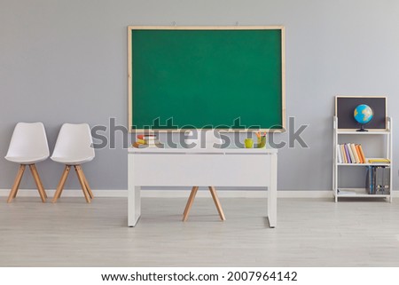 Interior of a modern light empty school classroom with a clean green blackboard, teacher's desk, chairs, shelf, books and earth globe. Education and back to school concept