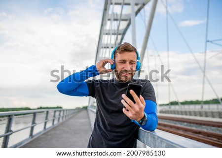 A man stands on a bridge and listens to music on headphones after training