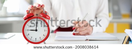 Businesswoman at her desk holds her hand on red alarm clock and makes entries in diary