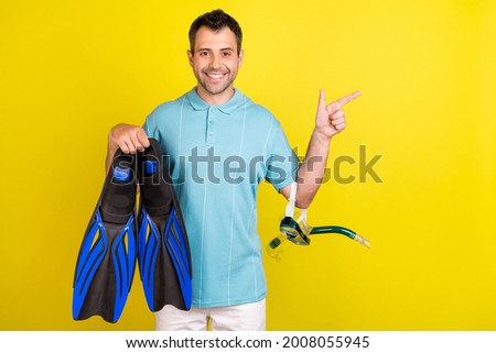 Photo of cute funny young man wear blue t-shirt holding flippers pointing empty space smiling isolated yellow color background