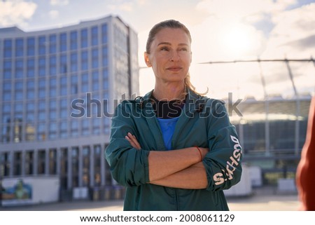 Professional middle aged sportswoman in sportswear looking away standing with arms crossed outdoors, ready for morning workout