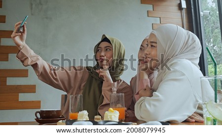 Group of young beautiful muslim women wearing hijab sitting near glass window in coffee shop, they are use smartphone to selfie with happiness