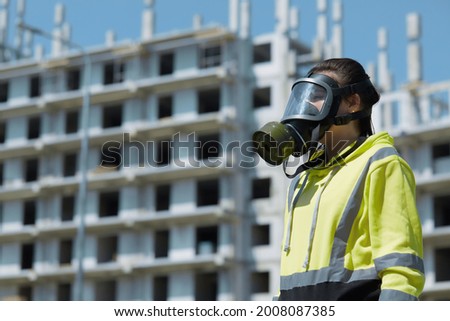 A close-up of a girl inspector in a gas mask for an environmental disaster is examined against the background of an unfinished industrial building.