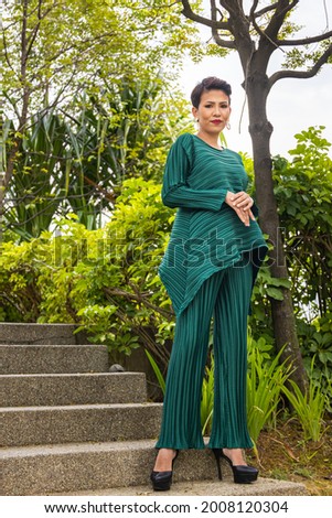 Young Asian woman leaning on a tree in a park. Young trendy model with black hair and green fashionable traditional local dress or female Baju Melayu. Fashion model on a summer day in a public park
