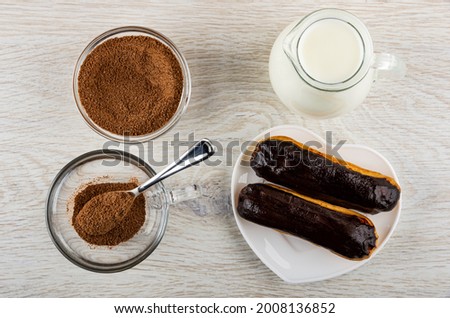 Cocoa powder with sugar in transparent bowl, pitcher with hot milk, teaspoon and cocoa in transparent cup, two eclairs with chocolate glaze in plate on wooden table. Top view