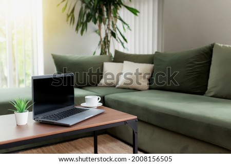 Work from home, workspace, desktop, remote work concept, gray laptop notebook, black empty blank screen on wooden table with white cup of coffee, green sofa, flowerpot. Stylish apartments living room