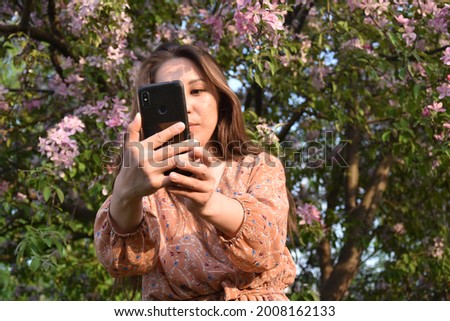 Soft focus of defocused young Asian woman in dress making selfie in blooming garden in sunlight 