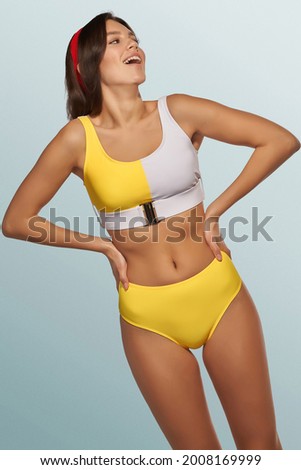  Slim lady is wearing two-piece swimsuit composed of gray and yellow bra with metal buckle and yellow high-waisted panties. Smiling girl with red headband is posing on the blue background.             Royalty-Free Stock Photo #2008169999
