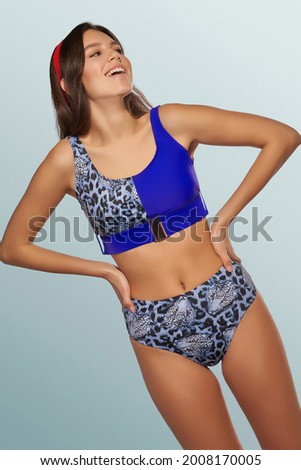 Slim lady is wearing blue two-piece swimsuit with leopard design composed of bra with metal buckle and high-waisted panties. Smiling girl with red headband is posing on the blue background.            Royalty-Free Stock Photo #2008170005