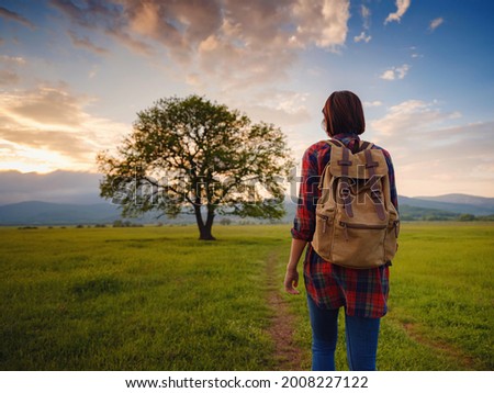 Asian traveler woman walk on the dirty road with sunshine and oak tree. In a plaid shirt and with a backpack. The end and the idea of adventure, travel, and discovery