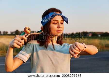 Outdoor photo of beautiful brunette woman in t shirt and hairband looking at her wristwatch while skateboarding, holding longboard over shoulders, has concentrated facial expression.