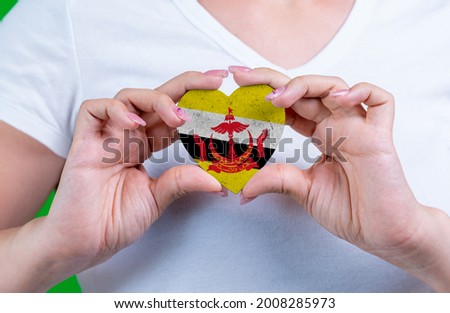Woman in white t-shirt holds a heart in the form flag Brunei on her chest. Photo for a postcard on Flag Day, Independence Day, travel, patriotism, other holiday