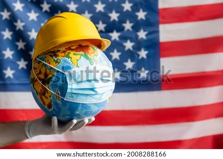 cropped view of man in latex glove holding globe with hardhat and medical mask near blurred usa flag, labor day concept