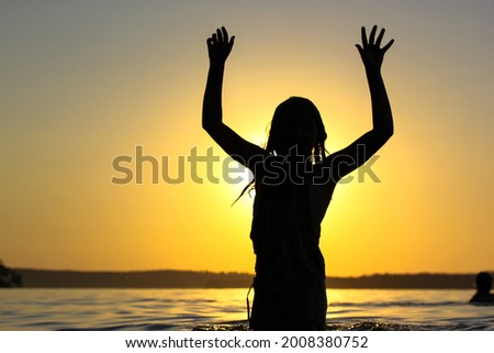 Silhouette of happy teenage girl standing a confident pose in a river, pond, lake water at sunset with hands up in the air, fingers keeps apart. Summer vacations, holidays on a coast. Happy childhood.