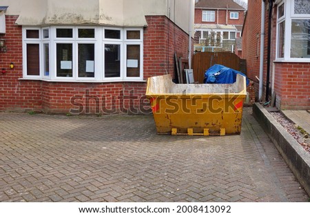 Big industrial yellow rubbish skip on driveway. Space to add text on surrounding background, front, side area of metal bin, dirty floor and brick houses. Great for renovate, moving, clearance concept.