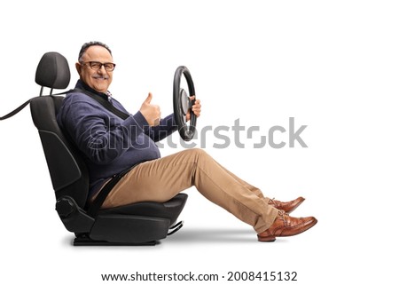 Cheerful mature man sitting in a car seat, holding a steering wheel and showing thumbs up isolated on white background