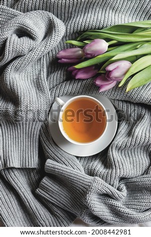 Spring composition flat lay white cup of tea, beautiful bouquet of violet tulips, grey sweater, concept of spring and comfort. Women’s day card, poster, wallpaper. Still life morning photography.
